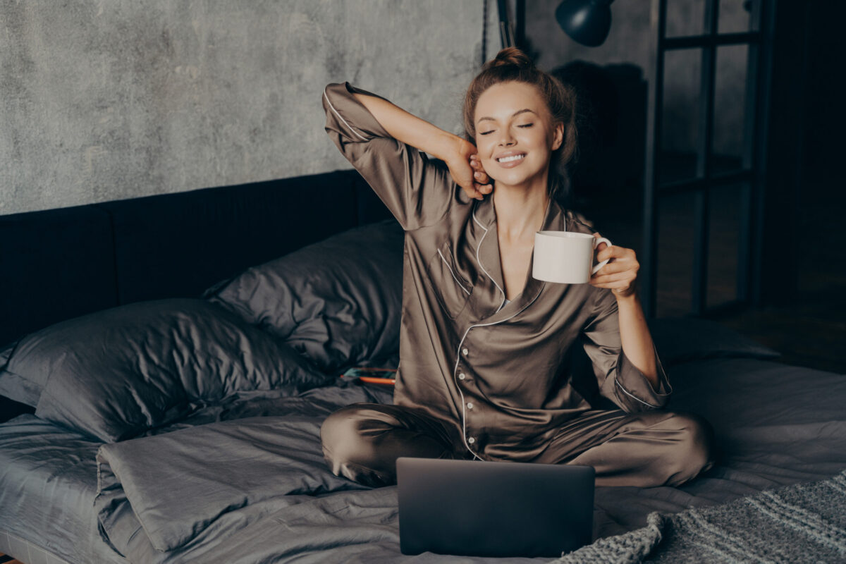 Relaxed female freelancer having cup of coffee stright after waking up in bed while checking new messages and emails on laptop, dressed in pajama. Woman with closed eyes ready to start remote work