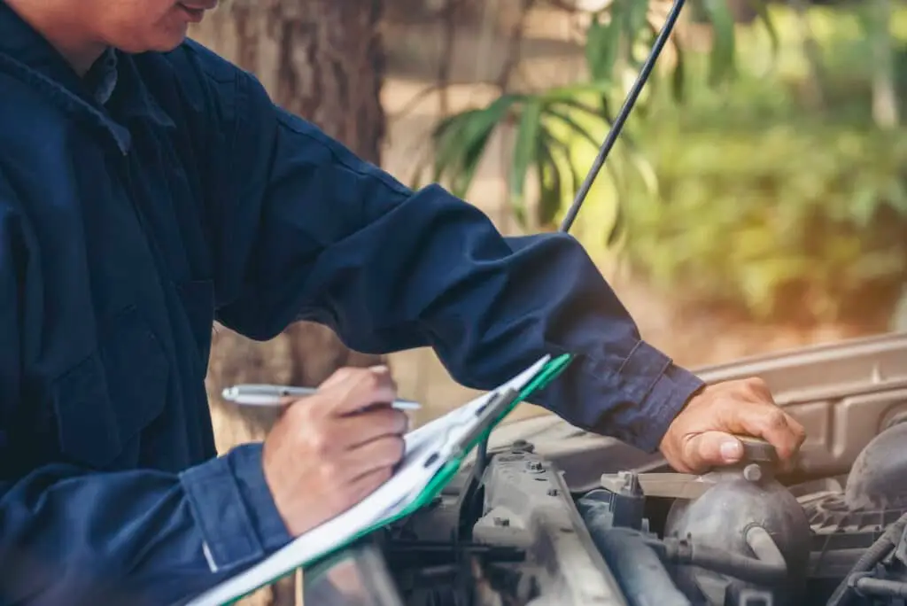 A person in a dark blue coverall inspecting the engine of a vehicle while taking notes on a clipboard