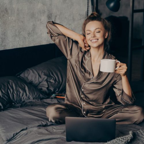 Relaxed female freelancer having cup of coffee stright after waking up in bed while checking new messages and emails on laptop, dressed in pajama. Woman with closed eyes ready to start remote work