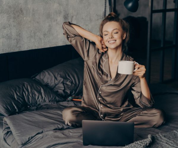 Relaxed female freelancer having cup of coffee stright after waking up in bed while checking new messages and emails on laptop, dressed in pajama. Woman with closed eyes ready to start remote work