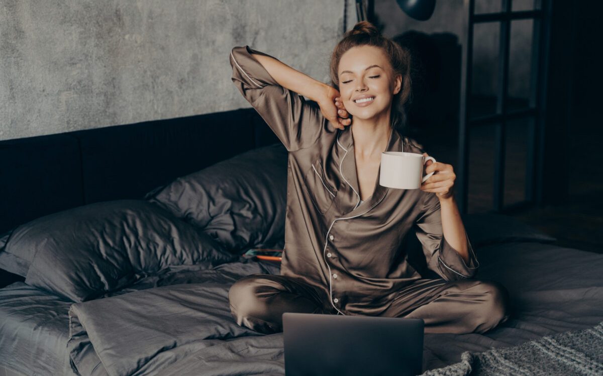 Relaxed female freelancer having cup of coffee stright after waking up in bed while checking new messages and emails on laptop, dressed in pajama. Woman with closed eyes ready to start remote work